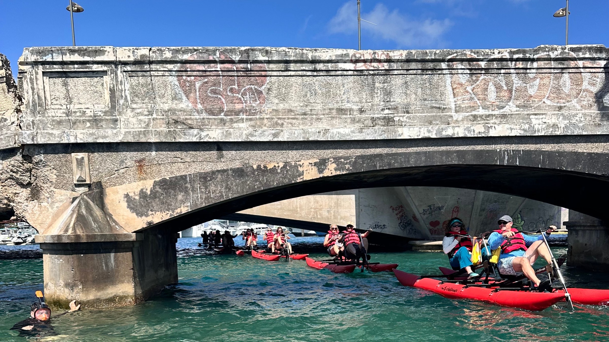 Two people riding a red waterbike on calm blue waters near a historic fort in San Juan on a sunny day.