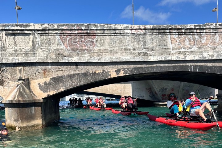 Two people riding a red waterbike on calm blue waters near a historic fort in San Juan on a sunny day.