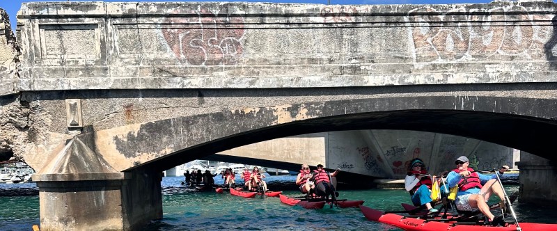 Two people riding a red waterbike on calm blue waters near a historic fort in San Juan on a sunny day.