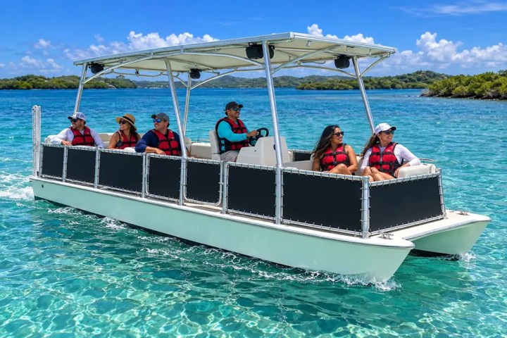 Pontoon boat with six passengers in life vests on clear blue water.