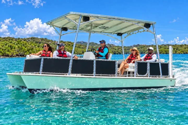 People on a pontoon boat wearing life vests, cruising through blue water near a wooded shoreline.