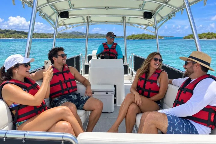 Group of five people on a boat wearing red life vests, smiling and enjoying a sunny day.